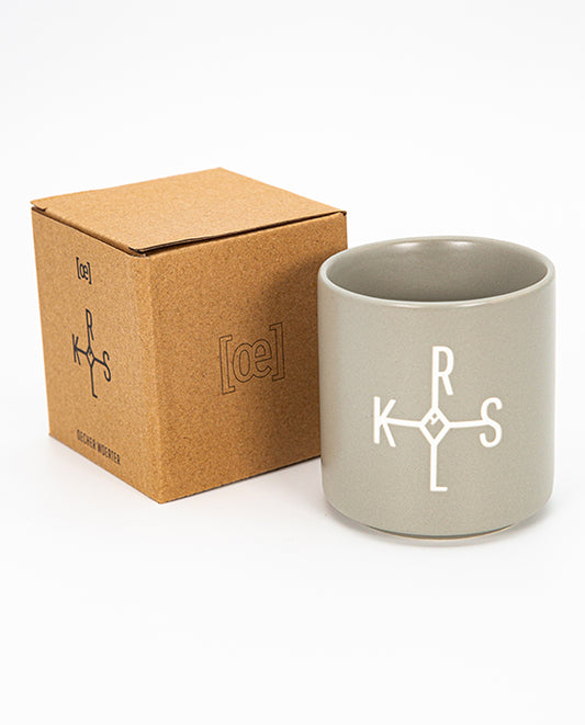 Grauer Keramikbecher mit matter Oberfläche und weißem grafischen Aufdruck „K R L S“; daneben braune Produktverpackung aus Karton mit passendem Aufdruck.