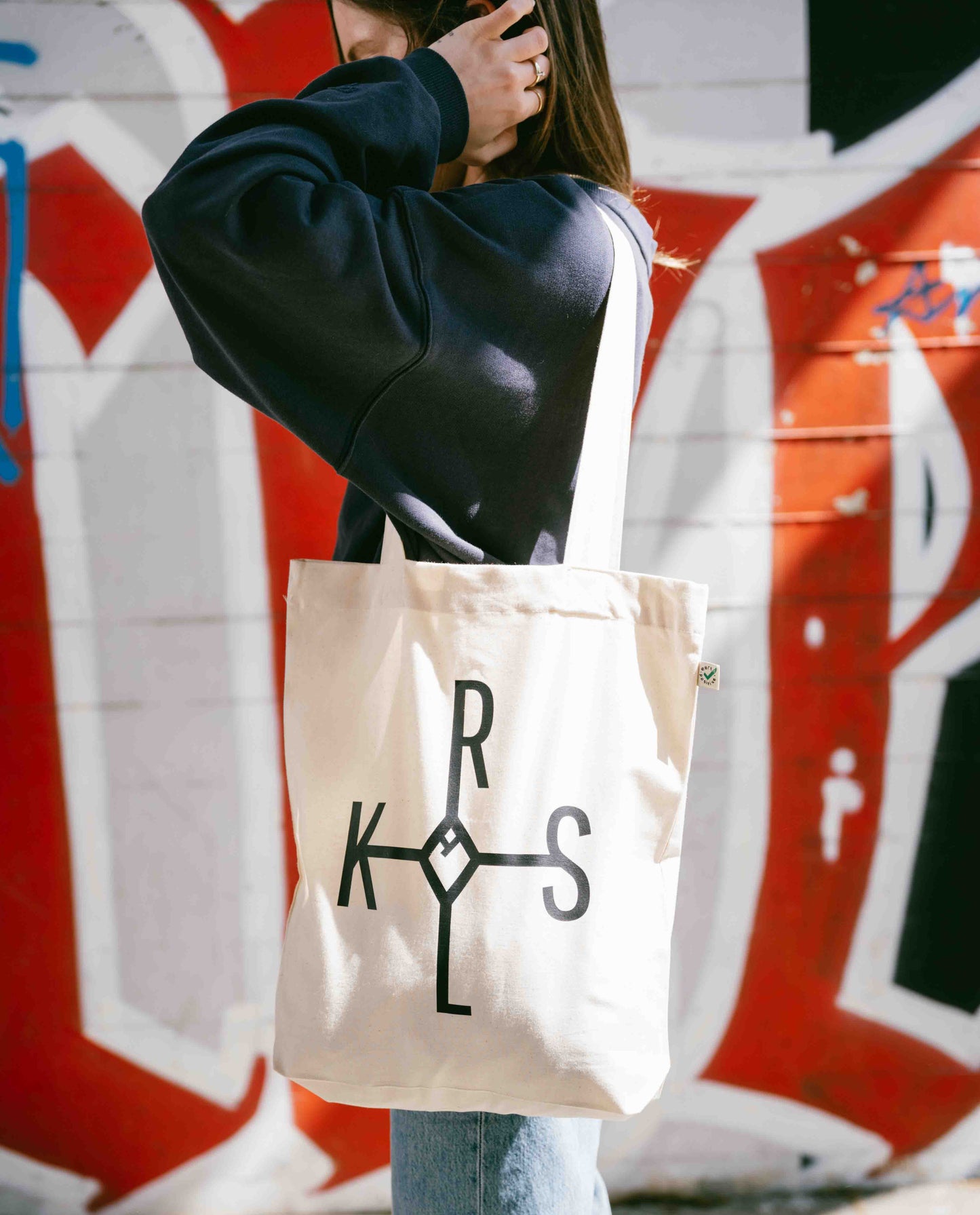 Seitliche Ansicht einer Frau mit beiger KRLS-Stofftasche vor rot-weißer Graffiti-Wand, trägt dunkelblauen Hoodie und Jeans.