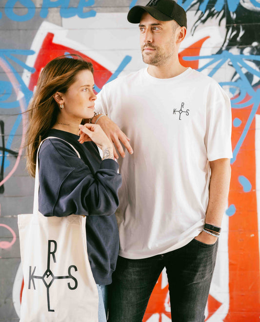 Zwei Personen in urbaner Umgebung tragen KRLS-Merch: ein weißes T-Shirt und ein Jutebeutel mit dem KRLS-Siegel