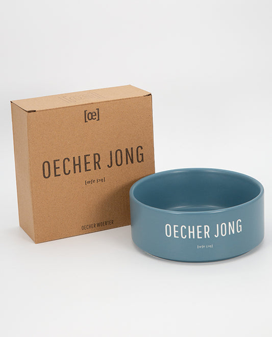 Blauer Keramiknapf mit weißem Aufdruck „OECHER JONG“ präsentiert neben einer braunen, bedruckten Kartonverpackung mit gleichem Schriftzug. Das Produkt steht vor einem neutralen weißen Hintergrund.