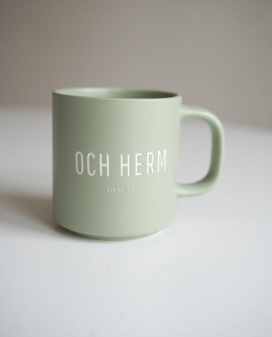 Matt-salbeigrüne Keramiktasse mit weißer Aufschrift „OCH HERM“ und kleiner Lautschrift darunter, auf weißer Fläche vor neutralem Hintergrund.