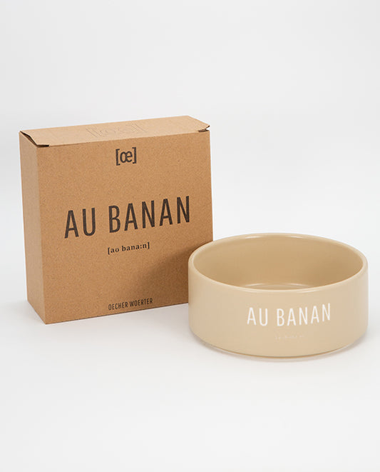 Beigefarbener Keramiknapf mit weißem Schriftzug „AU BANAN“ und passender brauner Produktverpackung im Hintergrund, auf hellem Hintergrund fotografiert.