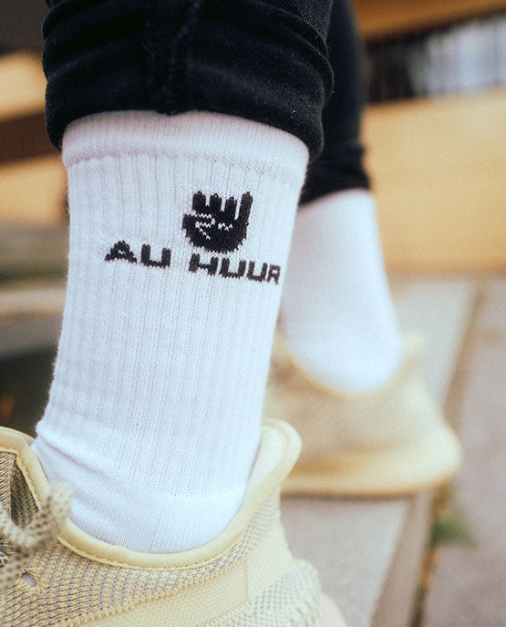 Person trägt weiße AU HUUR-Socken mit schwarzem Logo und Klenkes-Motiv, kombiniert mit beigen Sneakern und schwarzer Hose, fotografiert im urbanen Outdoor-Setting.