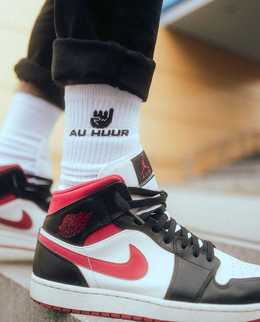 Weiße AU HUUR-Socken mit schwarzem Logo und Handzeichen, getragen mit Nike Air Jordan Sneakern in Schwarz-Weiß-Rot, lässiger Streetstyle-Look im urbanen Umfeld.
