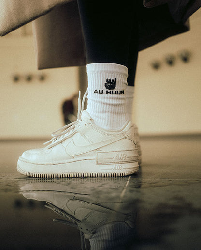 Weiße AU HUUR-Socken mit schwarzem Logo und Handzeichen, getragen mit weißen Nike Air Force 1 Shadow Sneakern und schwarzer Leggings, urbaner Look mit reflektierendem Boden.