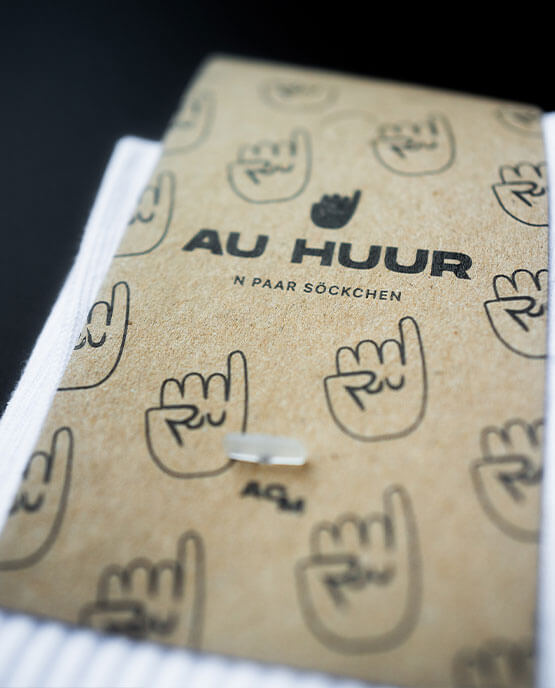 Detailansicht der Verpackung der weißen „AU HUUR“-Socken mit braunem Karton-Label, schwarzem Markenlogo und mehreren Klenkes Symbolen