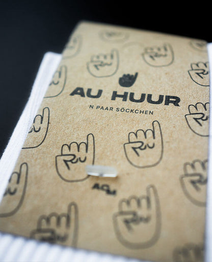 Detailansicht der Verpackung der weißen „AU HUUR“-Socken mit braunem Karton-Label, schwarzem Markenlogo und mehreren Klenkes Symbolen