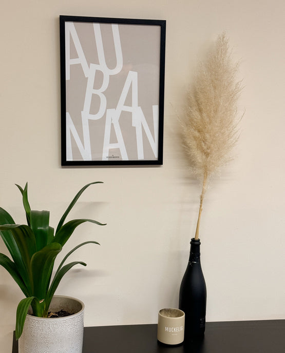 Beigefarbenes Poster mit dem Schriftzug „AU BANAN“ in großen weißen Buchstaben, eingerahmt in Schwarz. Daneben Pampasgras in einer schwarzen Flasche, ein Keramikbecher mit „MUCKELIG“-Aufdruck und eine grüne Pflanze in einem hellen Topf.