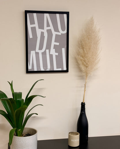 Poster mit dem Schriftzug „HAU DE MULL“ in weißer Typografie auf grauem Hintergrund, in schwarzem Rahmen, daneben Pampasgras in Flasche, Zimmerpflanze und beige Tasse mit der Aufschrift „MUCKELIG“ auf dunklem Tisch.