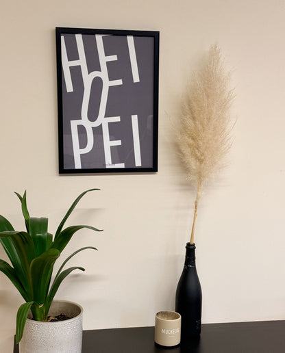 Wandposter mit dem Text „HEIOPEI“ in großformatiger, weißer Schrift auf anthrazitfarbenem Hintergrund. Gerahmt in Schwarz, daneben Pampasgras in dunkler Flasche, beige Tasse mit „MUCKELIG“-Aufdruck und grüne Zimmerpflanze im hellen Übertopf.