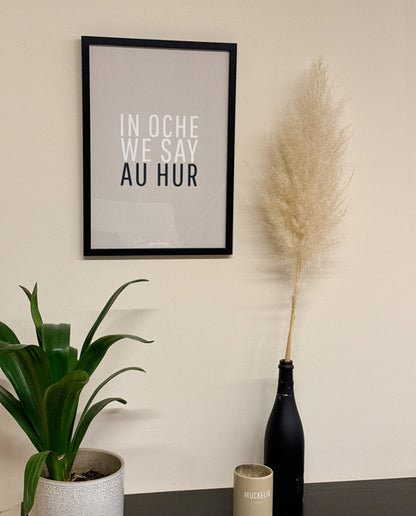 Poster mit dem Schriftzug „IN OCHE WE SAY AU HUR“ in schwarzem Rahmen an beiger Wand, daneben Pampasgras in schwarzer Flasche, grüne Pflanze und beige Kerze mit Aufschrift „MUCKELIG“ auf schwarzem Tisch.