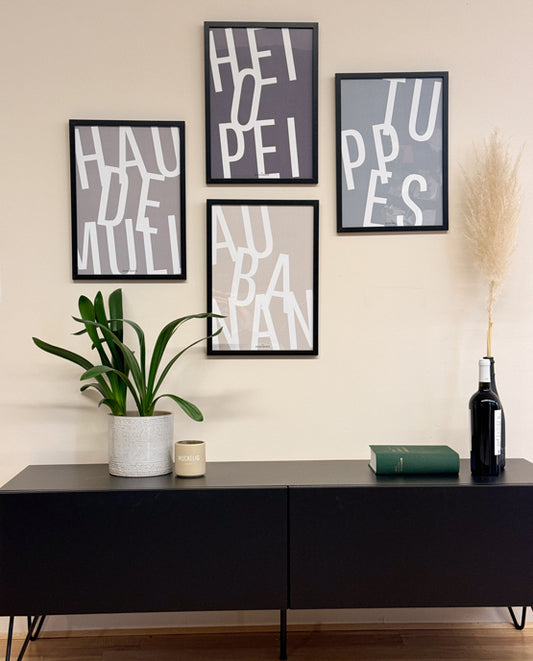 Poster mit Aachener Ausdrücken („HAU DE MULL“, „HEIOPEI“, „TUPPES“, „AU BANAN“) an einer beigefarbenen Wand. Darunter ein schwarzes Sideboard mit Zimmerpflanze im Topf, Becher mit „MUCKELIG“-Schriftzug, grünem Buch und schwarzer Flasche mit Pampasgras.