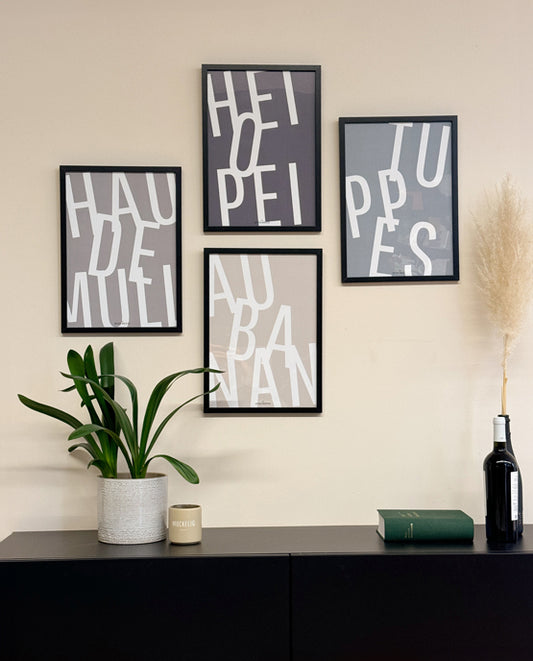Vier moderne Poster im Aachener Dialekt an einer beigefarbenen Wand: „HAU DE MULL“, „HEIOPEI“, „TUPPES“ und „AU BANAN“, jeweils in großen weißen Buchstaben auf unterschiedlichen Hintergründen. Darunter ein Sideboard mit einer grünen Pflanze, einem Becher mit „MUCKELIG“-Aufdruck, einem grünen Buch und einer schwarzen Flasche mit Pampasgras.