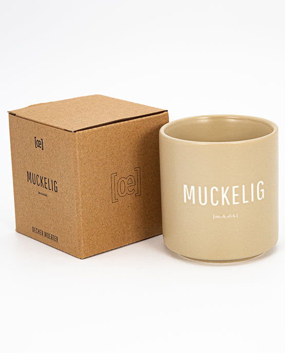 Beigefarbener Keramikbecher mit weißem Aufdruck „MUCKELIG“ und kleiner Lautschrift darunter, daneben die passende Verpackung in braun.