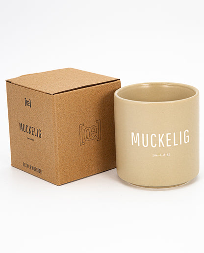 Beigefarbener Keramikbecher mit weißem Aufdruck „MUCKELIG“ und kleiner Lautschrift darunter, daneben die passende Verpackung in braun.
