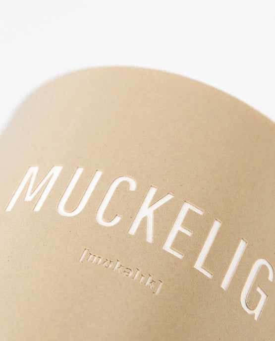 Nahaufnahme eines beigefarbenen Keramikbechers mit weißem Aufdruck „MUCKELIG“ und kleiner Lautschrift darunter.