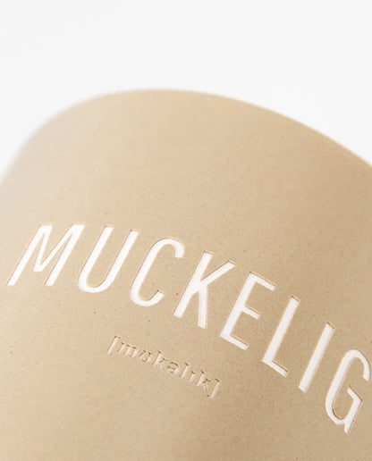 Nahaufnahme eines beigefarbenen Keramikbechers mit weißem Aufdruck „MUCKELIG“ und kleiner Lautschrift darunter.