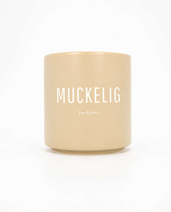 Beigefarbener Keramikbecher mit matter Oberfläche und weißem Aufdruck „MUCKELIG“ sowie kleinerer Lautschrift darunter; schlichtes, modernes Design.