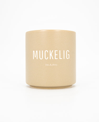 Beigefarbener Keramikbecher mit matter Oberfläche und weißem Aufdruck „MUCKELIG“ sowie kleinerer Lautschrift darunter; schlichtes, modernes Design.