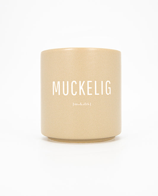 Beigefarbener Keramikbecher mit matter Oberfläche und weißem Aufdruck „MUCKELIG“ sowie kleinerer Lautschrift darunter; schlichtes, modernes Design.