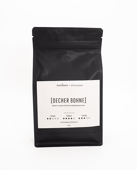 Mattschwarze Kaffeepackung mit weißem Etikett der Sorte „OECHER BOHNE“ von Oecher Woerter x d’Anastassio. Auf dem Etikett sind Geschmacksnoten (schokoladig, kräftig, rund) sowie Angaben zur Süße, Stärke und Crema abgebildet.