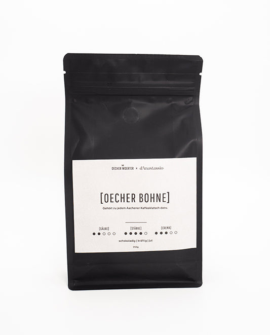 Mattschwarze Kaffeepackung mit weißem Etikett der Sorte „OECHER BOHNE“ von Oecher Woerter x d’Anastassio. Auf dem Etikett sind Geschmacksnoten (schokoladig, kräftig, rund) sowie Angaben zur Süße, Stärke und Crema abgebildet.