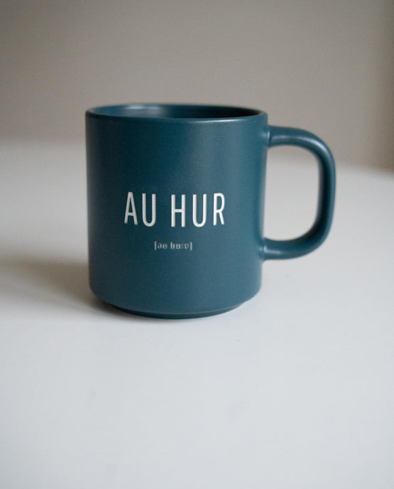 Petrolfarbene Keramiktasse mit weißem Aufdruck „AU HUR“ und phonetischer Schreibweise darunter. Schlichte, moderne Form mit rundem Henkel, auf weißem Hintergrund fotografiert.
