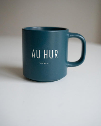 Petrolfarbene Keramiktasse mit weißem Aufdruck „AU HUR“ und phonetischer Schreibweise darunter. Schlichte, moderne Form mit rundem Henkel, auf weißem Hintergrund fotografiert.