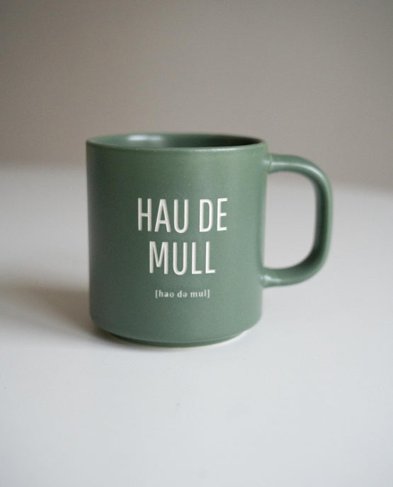 Grüne Keramiktasse mit weißer Aufschrift „HAU DE MULL“ und darunter phonetischer Schreibweise auf weißer Oberfläche platziert.