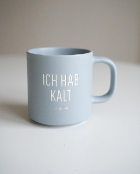 Hellblaue Keramiktasse mit weißer Aufschrift „ICH HAB KALT“ und kleiner Lautschrift darunter, aufgestellt auf weißer Fläche vor neutralem Hintergrund.