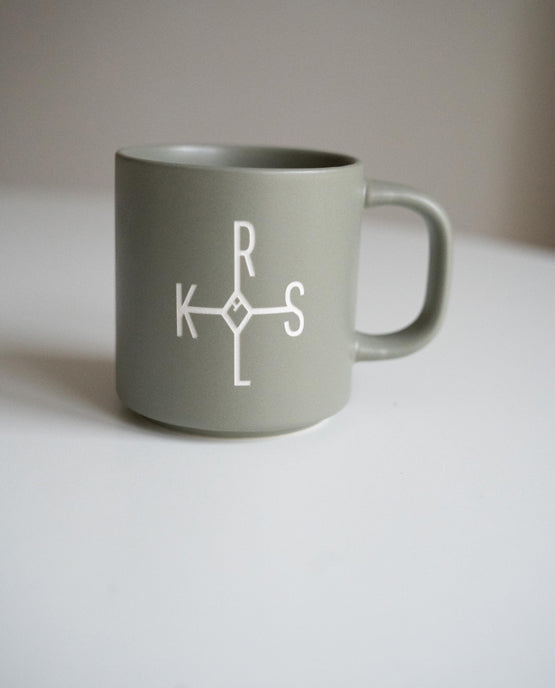 Graue Keramiktasse mit weißem Karolus-Logo bestehend aus den Buchstaben „K“, „R“, „L“ und „S“ auf weißer Fläche vor neutralem Hintergrund.