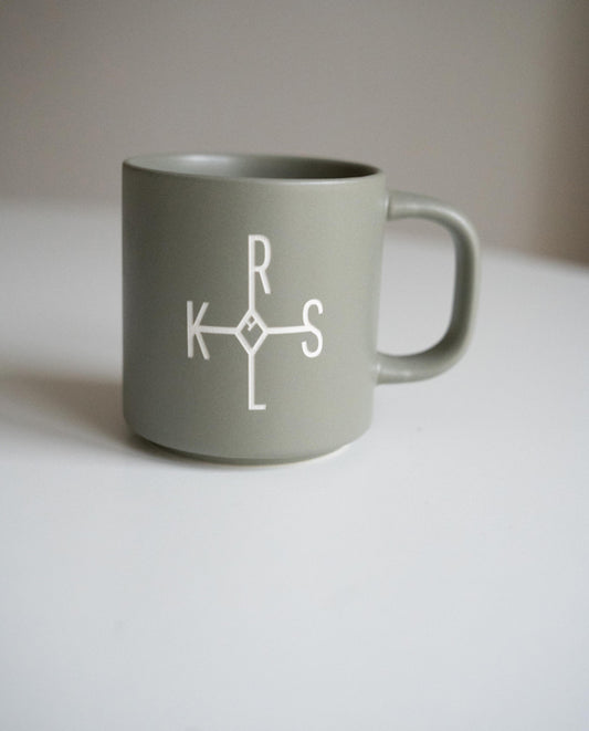 Graue Keramiktasse mit weißem Karolus-Logo bestehend aus den Buchstaben „K“, „R“, „L“ und „S“ auf weißer Fläche vor neutralem Hintergrund.