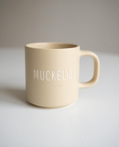 Matt beige Keramiktasse mit weißer Aufschrift „MUCKELIG“ und kleiner Lautschrift darunter, aufgestellt auf weißer Fläche vor neutralem Hintergrund.