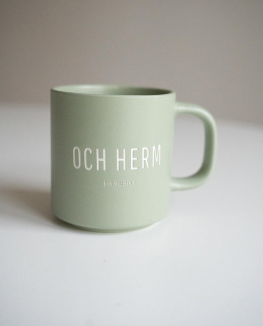 Matt-salbeigrüne Keramiktasse mit weißer Aufschrift „OCH HERM“ und kleiner Lautschrift darunter, auf weißer Fläche vor neutralem Hintergrund.