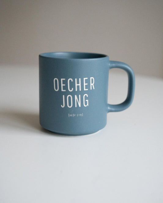 Blaue Keramiktasse mit weißer Aufschrift „OECHER JONG“ und kleiner Lautschrift darunter, auf weißer Fläche vor neutralem Hintergrund.