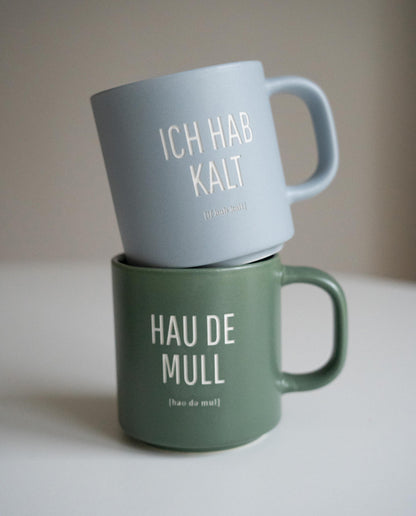 Zwei Keramiktassen mit Aachener Sprüchen, übereinandergestapelt auf weißer Oberfläche: oben eine hellblaue Tasse mit der Aufschrift „ICH HAB KALT“, unten eine grüne Tasse mit der Aufschrift „HAU DE MULL“.