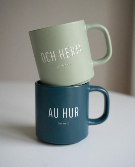 Zwei Keramiktassen übereinandergestapelt auf weißer Oberfläche. Die obere Tasse ist hellgrün mit weißer Aufschrift „OCH HERM“ und phonetischer Schreibweise. Die untere Tasse ist dunkelblau mit der Aufschrift „AU HUR“ und phonetischer Schreibweise.