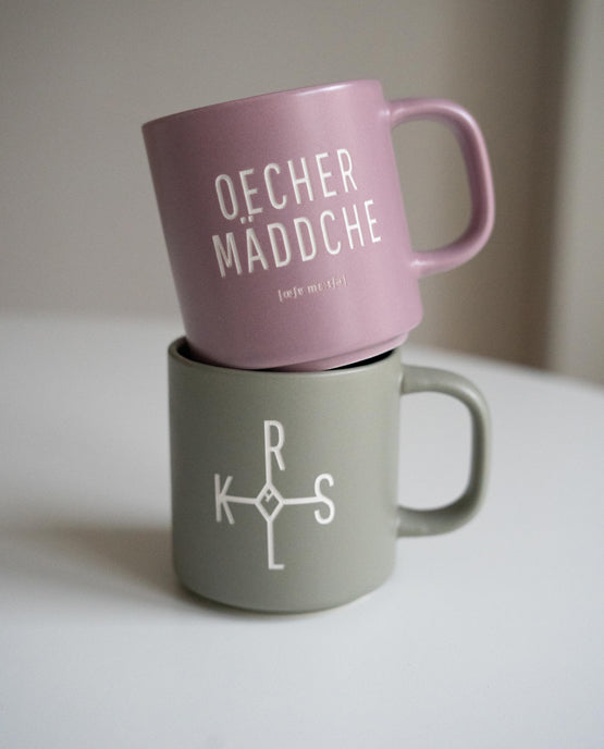 Zwei Keramiktassen übereinandergestapelt auf weißer Oberfläche. Die obere Tasse ist matt-rosa mit weißer Aufschrift „OECHER MÄDDCHE“ und phonetischer Schreibweise. Die untere Tasse ist grau mit dem weißen Karolus-Symbol aus den Buchstaben „K“, „R“,„L“, „S“ und die durch Linien verbunden sind.