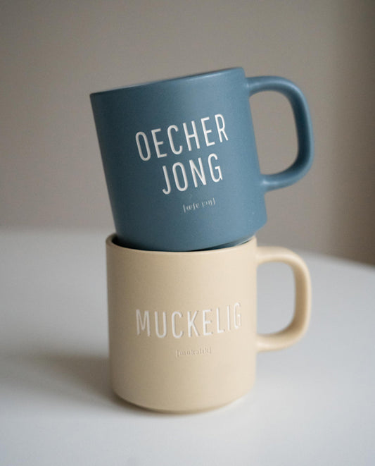 Zwei Keramiktassen übereinandergestapelt auf weißer Oberfläche. Die obere Tasse ist mattblau mit weißer Aufschrift „OECHER JONG“ und phonetischer Schreibweise. Die untere Tasse ist beige mit der Aufschrift „MUCKELIG“ und phonetischer Schreibweise.