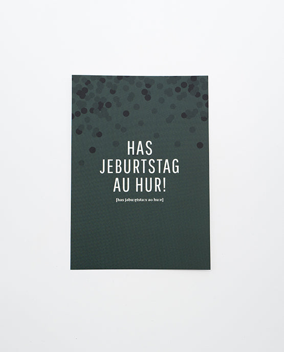 Dunkelgrüne Postkarte mit schwarzem Konfettimuster im oberen Bereich und weißer Aufschrift „HAS JEBURTSTAG AU HUR!“ in Großbuchstaben, darunter phonetische Schreibweise in kleinerer Schrift