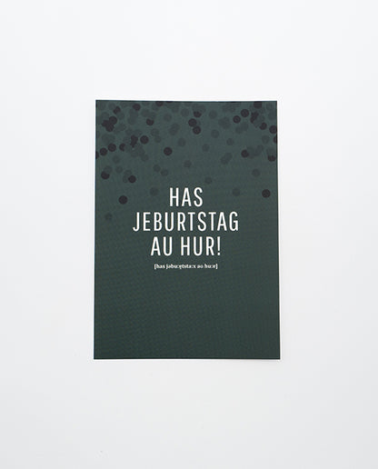 Dunkelgrüne Postkarte mit schwarzem Konfettimuster im oberen Bereich und weißer Aufschrift „HAS JEBURTSTAG AU HUR!“ in Großbuchstaben, darunter phonetische Schreibweise in kleinerer Schrift