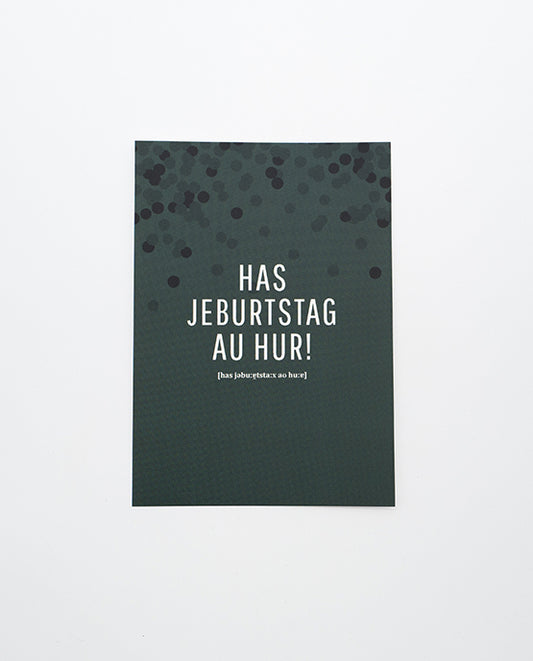 Dunkelgrüne Postkarte mit schwarzem Konfettimuster im oberen Bereich und weißer Aufschrift „HAS JEBURTSTAG AU HUR!“ in Großbuchstaben, darunter phonetische Schreibweise in kleinerer Schrift