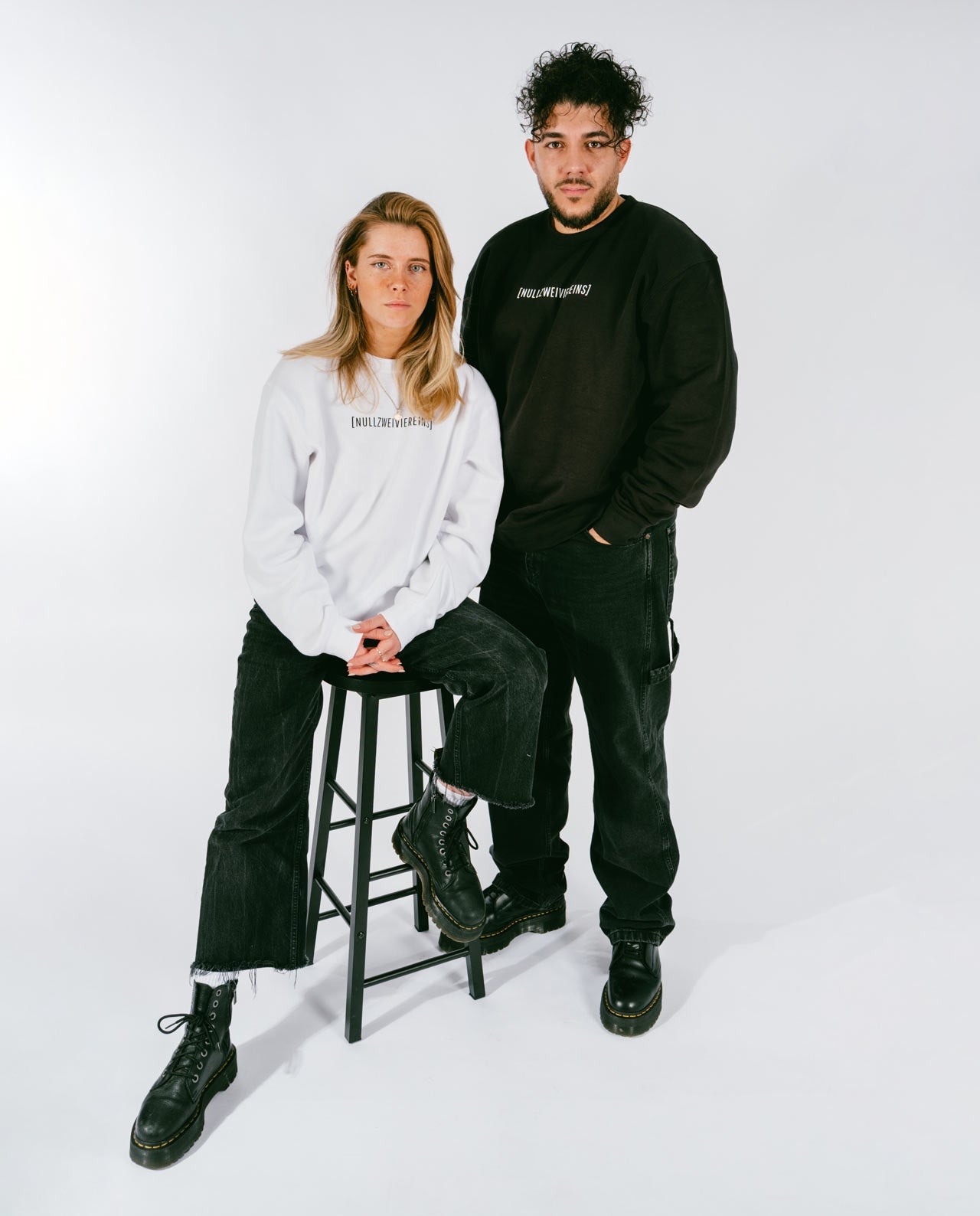 Frau sitzt auf einem schwarzen Hocker und trägt ein weißes Sweatshirt mit schwarzem Schriftzug „[NULLZWEIVIEREINS]“, schwarzer Jeans und schwarzen Schnürboots. Neben ihr steht ein Mann in einem schwarzen Sweatshirt mit weißem Schriftzug „[NULLZWEIVIEREINS]“, schwarzer Jeans und schwarzen Schnürboots. Beide vor weißem Hintergrund.