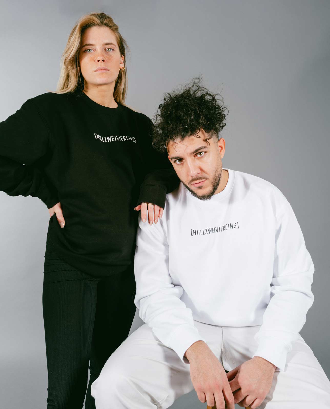 Frau steht in schwarzem Sweatshirt mit weißem Schriftzug „[NULLZWEIVIEREINS]“ und schwarzer Hose, während ein Mann davor sitzt und ein weißes Sweatshirt mit schwarzem Schriftzug „[NULLZWEIVIEREINS]“ sowie weiße Hose trägt. Beide posieren vor grauem Hintergrund.