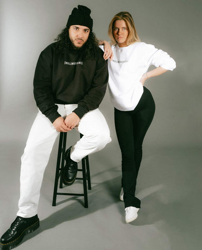 Mann mit schwarzer Mütze, schwarzem Sweatshirt mit weißem Schriftzug „[NULLZWEIVIEREINS]“ und weißer Hose sitzt auf einem schwarzen Hocker, neben einer Frau im weißen Sweatshirt mit schwarzem Schriftzug „[NULLZWEIVIEREINS]“ und schwarzer Hose. Beide posieren vor grauem Hintergrund.