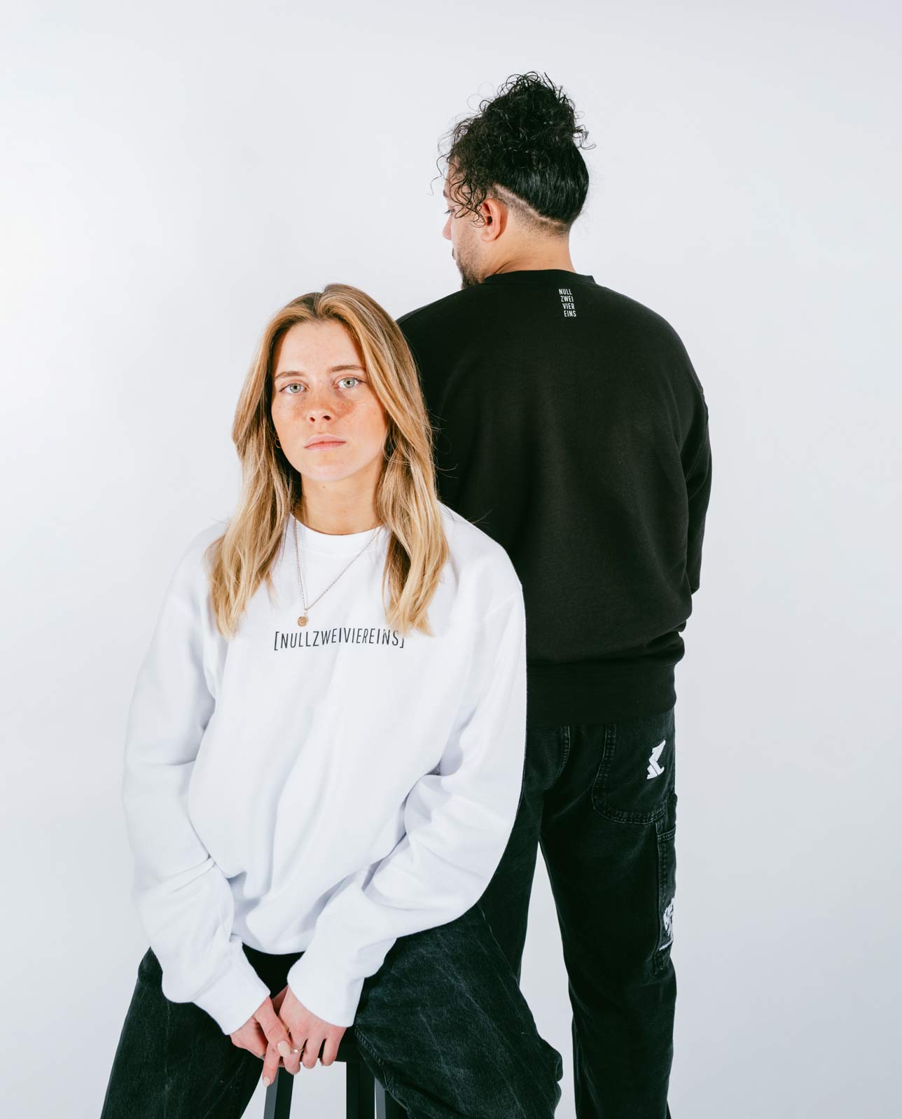 Frau sitzt auf einem Hocker und trägt ein weißes Sweatshirt mit schwarzem Schriftzug „[NULLZWEIVIEREINS]“, während ein Mann mit dem Rücken zur Kamera danebensteht und ein schwarzes Sweatshirt mit weißem Schriftzug im Nacken trägt. Beide vor weißem Hintergrund.
