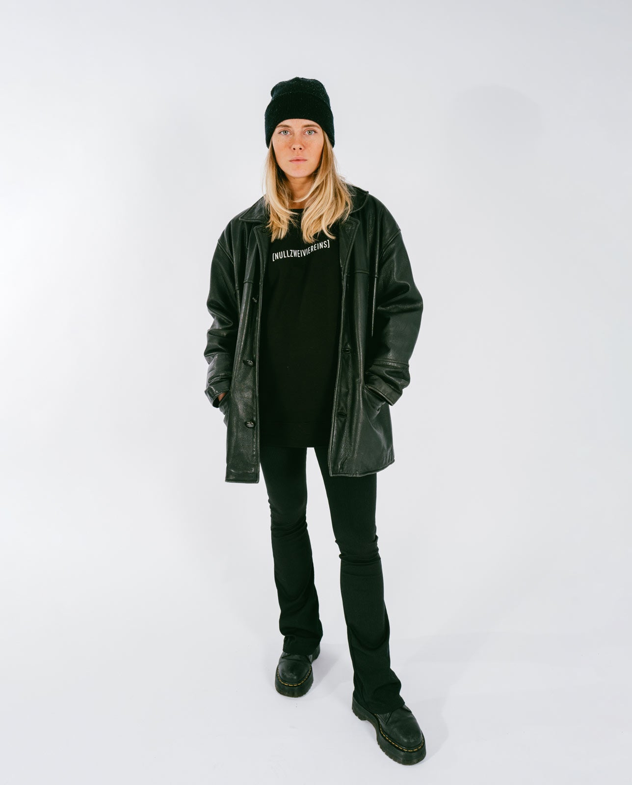 Frau steht vor weißem Hintergrund und trägt ein schwarzes T-Shirt mit weißem Schriftzug „[NULLZWEIVIEREINS]“, kombiniert mit schwarzer Lederjacke, schwarzer Mütze, schwarzer Hose und schwarzen Stiefeln.