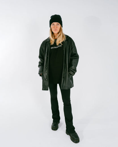 Frau steht vor weißem Hintergrund und trägt ein schwarzes T-Shirt mit weißem Schriftzug „[NULLZWEIVIEREINS]“, kombiniert mit schwarzer Lederjacke, schwarzer Mütze, schwarzer Hose und schwarzen Stiefeln.