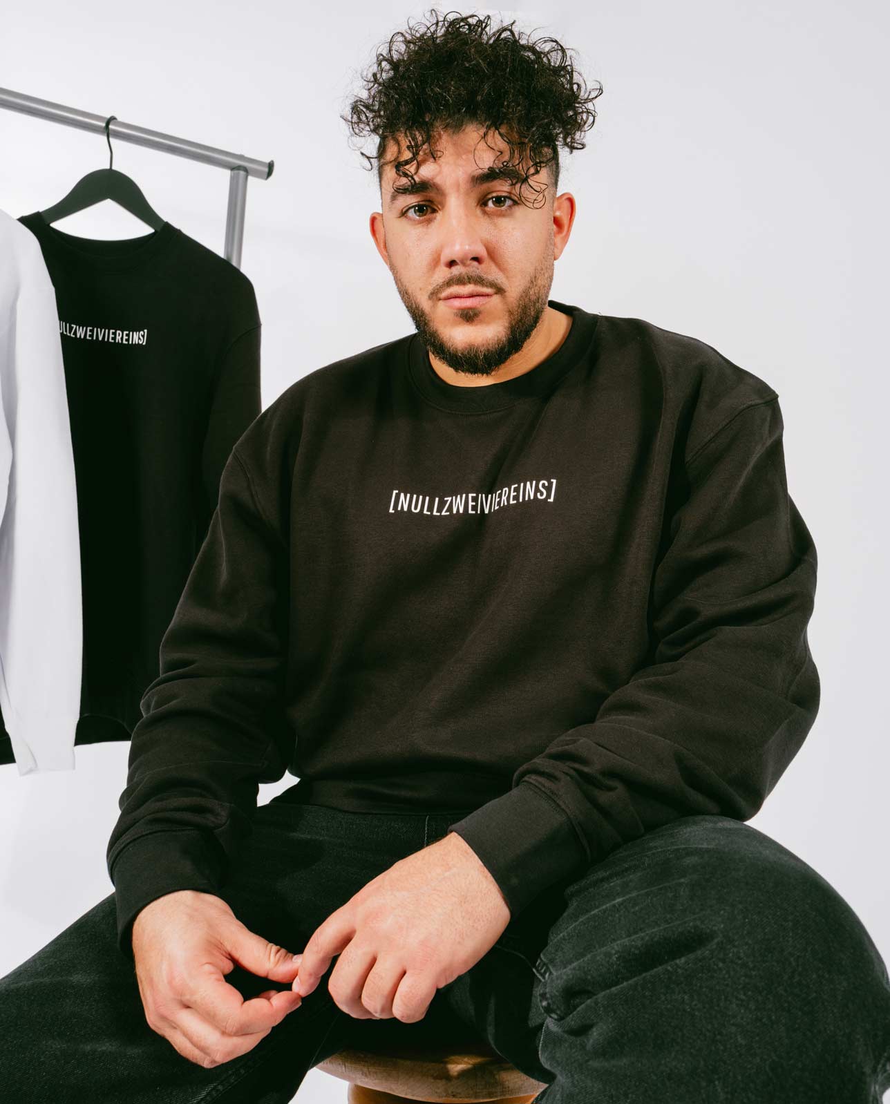 Mann sitzt auf einem Hocker vor weißem Hintergrund und trägt ein schwarzes Sweatshirt mit weißem Schriftzug „[NULLZWEIVIEREINS]“ auf der Brust, kombiniert mit schwarzen Jeans. Im Hintergrund hängt ein schwarzer und ein weißer Pullover auf einem Kleiderständer.