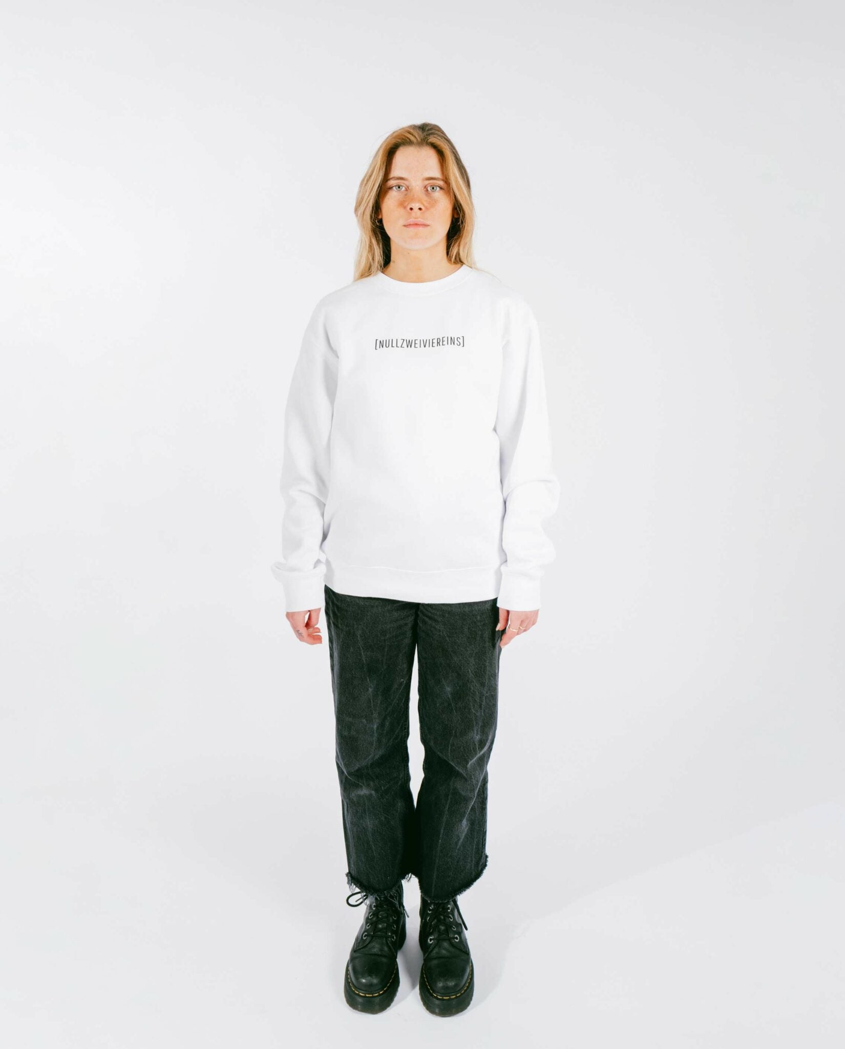 Frau steht frontal vor weißem Hintergrund und trägt einen weißen Sweatshirt mit schwarzem Schriftzug „[NULLZWEIVIEREINS]“ auf der Brust, kombiniert mit schwarzen, leicht verkürzten Jeans und schwarzen Schnürboots.
